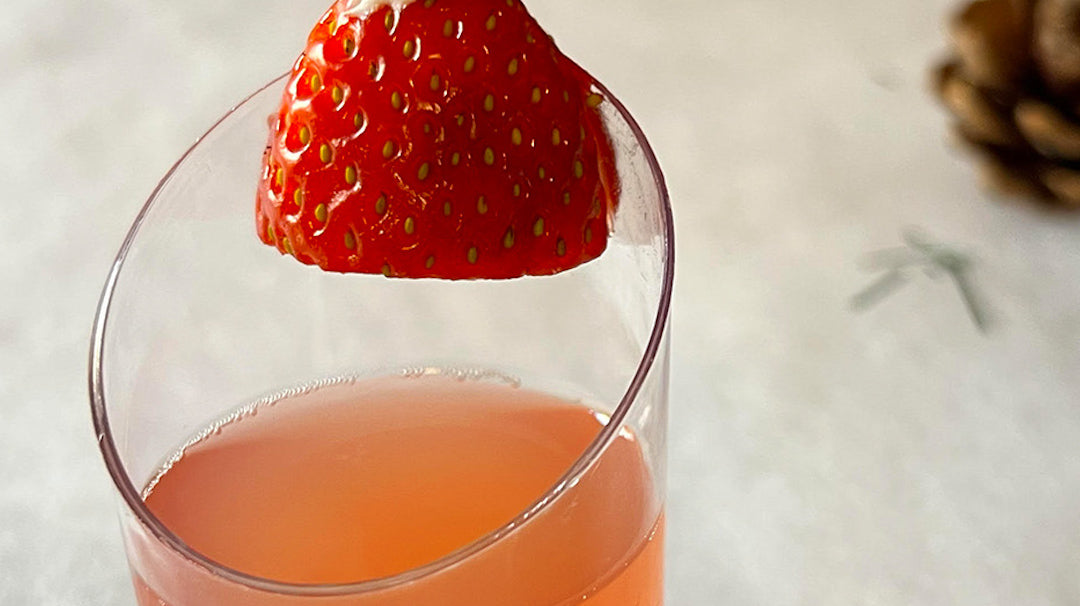 a cocktail shooter filled with red strawberry margarita mix and tequila. It has a cocktail garnish of a strawberry and dollop of Cool Whip to resemble the hat of Santa Claus